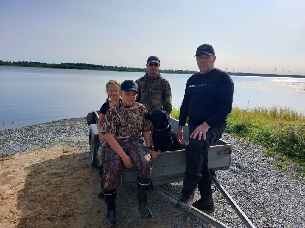 Luukas, Matilda ja Ari istuvat auton peräkärryllä järven rannalla. Mukana on myös neljäs henkilö, Sauli Sornikoski. Kaikki kuvan henkilöt ovat pukeutuneet metsästysasuihin.