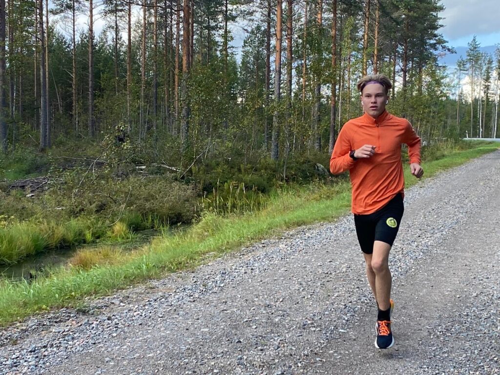 Mies juoksee metsäautotiellä. Miehellä on oranssi pusero.