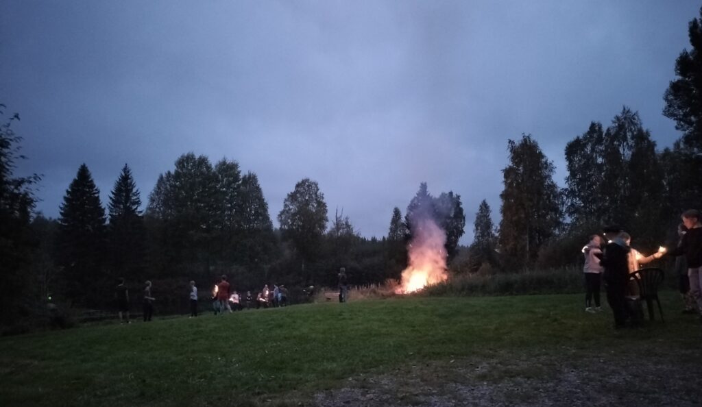 Pimenevä ilta, kokko palaa nurmikon laidassa. Ihmisiä seisoo pieninä ryppäinä siellä täällä. Lapset polttavat tähtisadetikkuja.