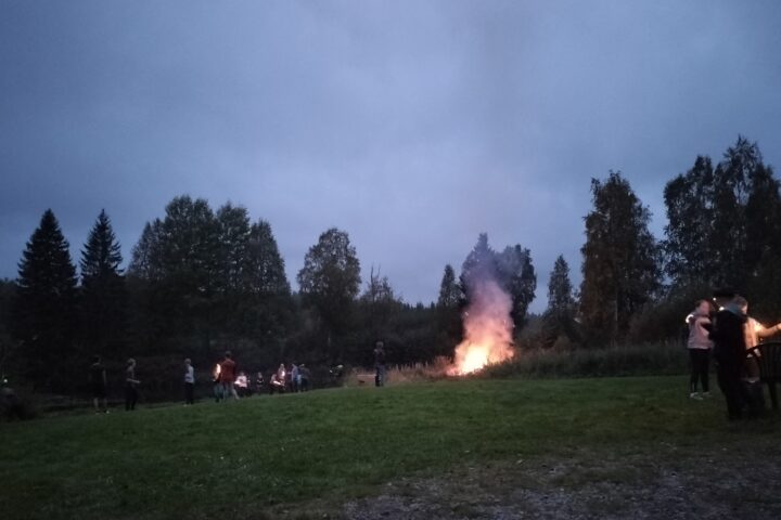 Pimenevä ilta, kokko palaa nurmikon laidassa. Ihmisiä seisoo pieninä ryppäinä siellä täällä. Lapset polttavat tähtisadetikkuja.